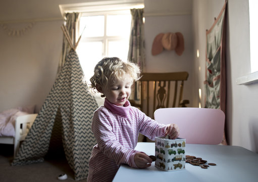Girl putting coins in piggy bank while standing by table at home