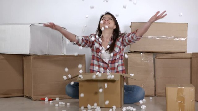 Happy woman unpacks moving cardboard boxes in a new apartment throwing polystyrene foam chips in the air
