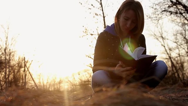 Beautiful young girl, sitting on a fallen autumn leaves in a park, reading a book, sunset light. Concept girl freelancer, working in a spring park.