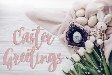 easter greetings text card sign on stylish easter eggs in nest with bunny on  fabric and tulips and willow on rustic white wooden background flat lay. space for text, top view.  happy easter