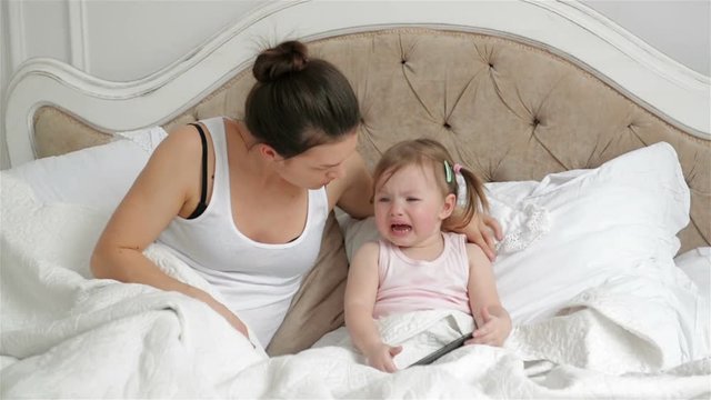 Cute Little Girl with Two Ponytails is Crying Sitting on the Bed. Mother is Trying to Calm Her Daughter in the Bedroom.