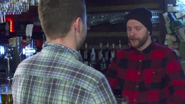 Two caucasian friends chatting at the pub. Attractive bearded guy in red checkered shirt and black hat gesturing by his hands. Brown haired man in gray clothes standing backwards near the bar counter
