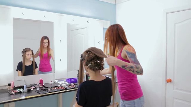 Professional hairdresser doing hairstyle for young pretty woman - combing hair and using spray. Beauty and haircare concept