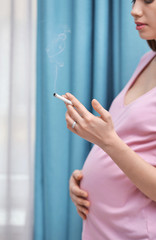 Pregnant woman smoking cigarette near window at home