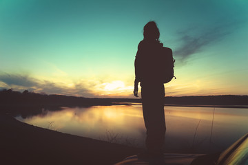 Young woman with backpack standing on car hood at sunset in front of a lake