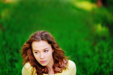 Naklejka premium Beautiful portrait of young pretty pensive brown-haired woman with wonderful hair on a blurry background of bright green grass with the sunlight, close-up.