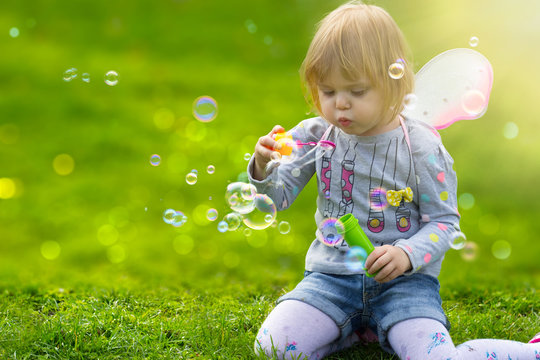 Toddler Girl With Butterfly Wings Having Fun In The Park, Blowing Soap Bubbles