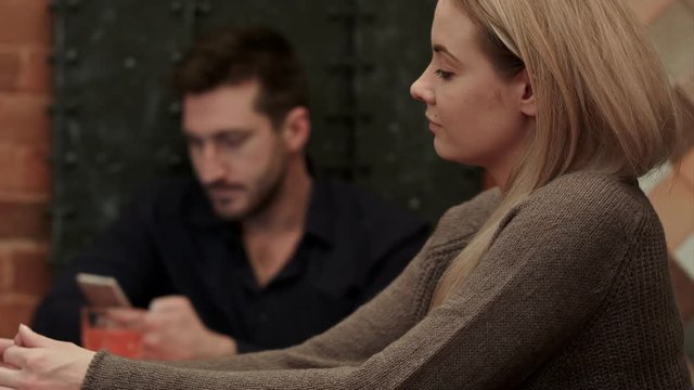 While woman stitting at the bar, man with cell phone behind looking at her