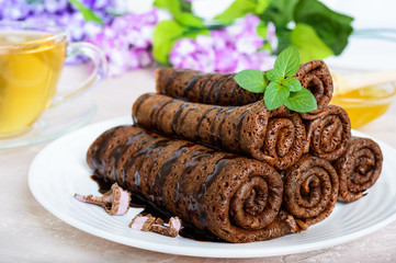Thin delicate chocolate pancakes, rolled, laid out in a pile on a white plate and a cup of herbal tea with honey
