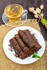 Thin delicate chocolate pancakes, rolled, laid out in a pile on a white plate and a cup of herbal tea with honey. Vertical view