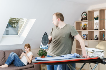 Obraz premium Young woman sitting at the sofa looking at smartphone while her husband is ironing shirt at home.