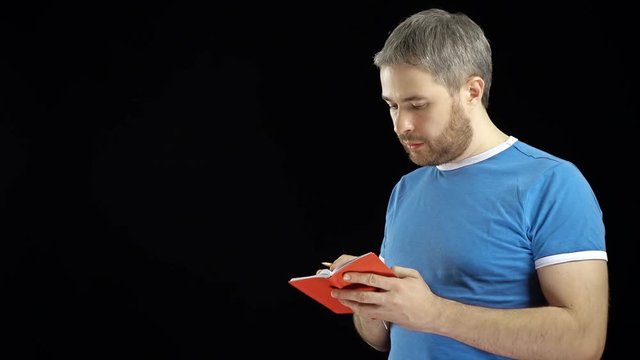 Handsome man in blue tshirt writing in his red notebook. Memo, search or contact concepts. Black background. 4K video