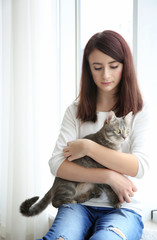 Beautiful young woman with cute cat sitting on window sill at home