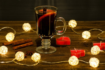 Mug of mulled wine with spices, candles in the shape of a heart on a wooden table, a garland of lanterns. Cinnamon sticks, anise