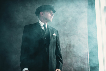 Retro 1920s english gangster wearing flat cap and suit. Looking out window.