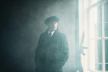 Retro 1920s english gangster wearing flat cap and coat. Standing near window.