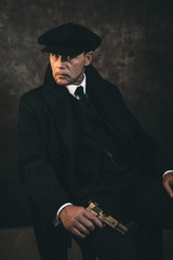 Retro 1920s english gangster with gun sitting on chair. Peaky blinders style.