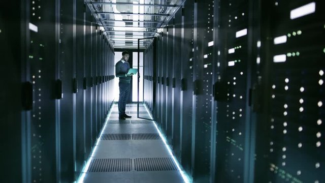 Male Server Engineer in Data Center. He Stands Before Open Door of Rack Server and Works on His Laptop. Shot on RED EPIC-W 8K Helium Cinema Camera.