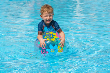 Fototapeta na wymiar Little boy in a swimming suit plays with an inflatable transparent ball in the spa pool. He looks to the side. Face view. Sunny day. Clear water.
