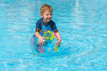 Little boy in a swimming suit plays with an inflatable transparent ball in the spa pool. He looks to the side. Face view. Sunny day. Clear water.