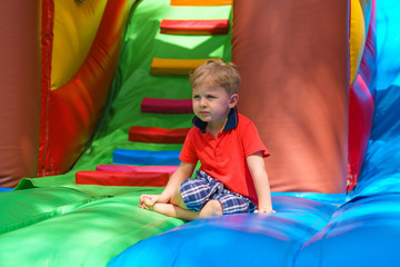 Fototapeta na wymiar Little boy is resting, sitting on a children's inflatable trampoline. He looks into the distance. Bright and motley colors. Sunny day.