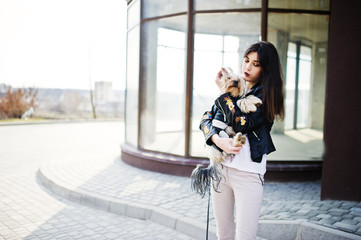 Brunette gypsy girl with yorkshire terrier dog posed against large windows house. Model wear on leather jacket and t-shirt with ornament, pants.