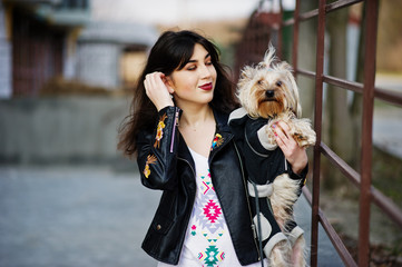 Brunette gypsy girl with yorkshire terrier dog posed against steel railings. Model wear on leather jacket and t-shirt with ornament, pants.