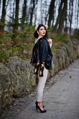 Brunette gypsy girl posed against stones on park. Model wear on leather jacket and t-shirt with ornament, pants and shoes with high heels.