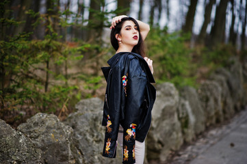 Naklejka na ściany i meble Brunette gypsy girl posed against stones on park. Model wear on leather jacket and t-shirt with ornament, pants.