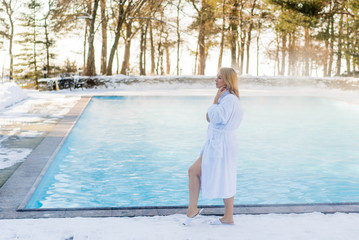 Young blonde woman in bathrobe near outdoor swimming pool at winter day