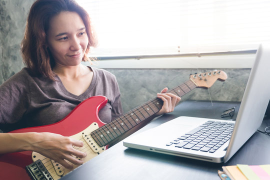Young woman playing guitar and using laptop on the table at home.