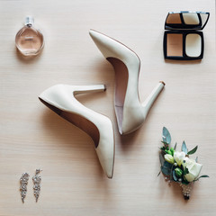 Powder set, rose boutonniere, parfume and crystal earrings lie around white shoes on high heels