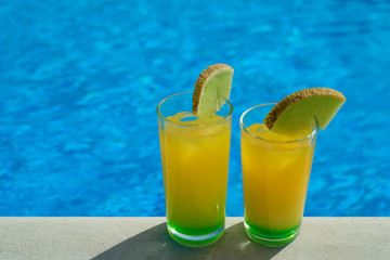 Glasses with yellow and green cocktail in front of a swimming pool with clear water. Close-up photo.
