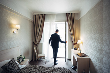 Groom in dark suit stands before bright balcony in the hotel room