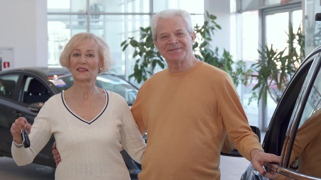 Attractive senior couple buying the car at the dealership. Gray caucasian man embracing his wife against background of new sedan. Aged blond woman showing car key near her husband