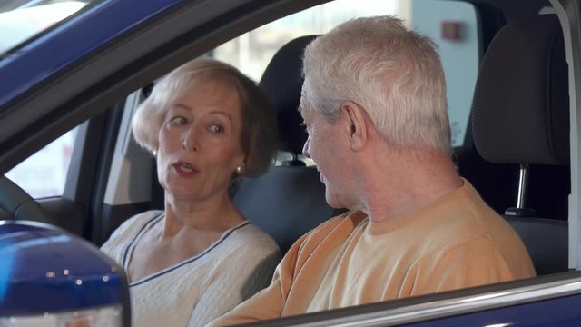 Attractive senior couple talking inside the car at the dealership. Aged caucasian man and woman sitting on the front seats of the vehicle. Brunette bearded guy discussing new car with his wife
