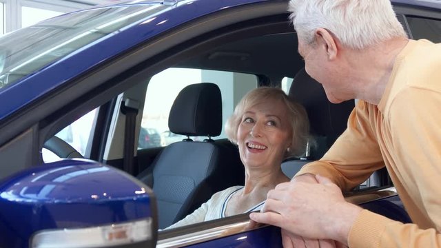 Attractive senior couple posing with car at the dealership. Aged gray man looking inside the purple crossover. Blond caucasian woman sitting on the driver's seat