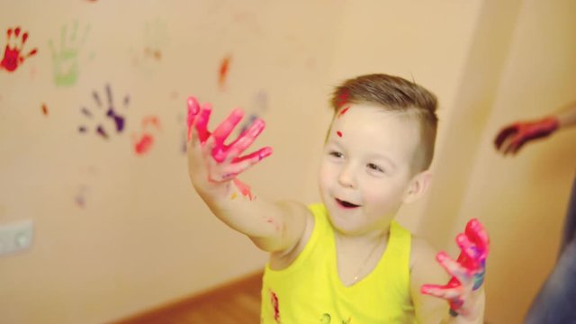 Cute boy showing his hands in paint. Happy little child making colorful handsprint on the wall with mother together.