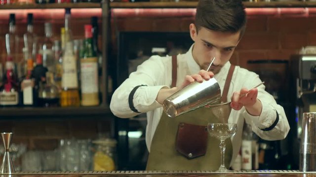 Young bearded barman prepare cocktail drink using two types of alcohol, sweet syrup in unusual glass, and decorating it