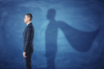 Businessman is standing in profile casting a shadow of the superman's cape on blue chalkboard...