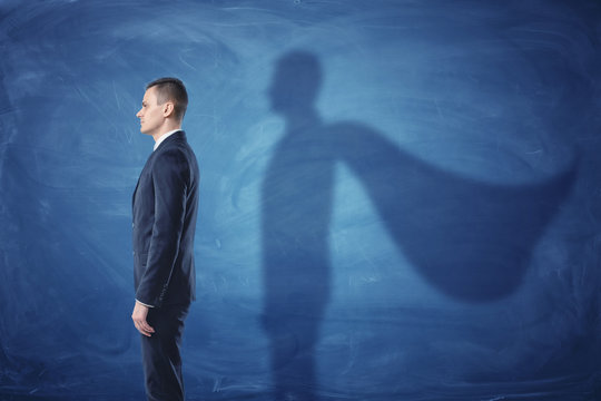 Businessman is standing in profile casting a shadow of the superman's cape on blue chalkboard background