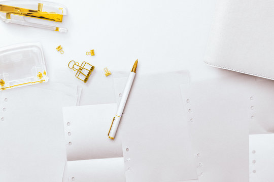 A desktop flatlay hero image, featuring white and gold items, a notepad, paper inserts and gold stationery accessories. Negative space to the top, on a plain white desk background.