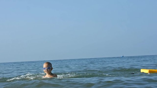 Happy smiling adorable little child enjoying blue sea surfboarding on bodyboard and jumping into the water on sky background on summer vacation in slow motion.