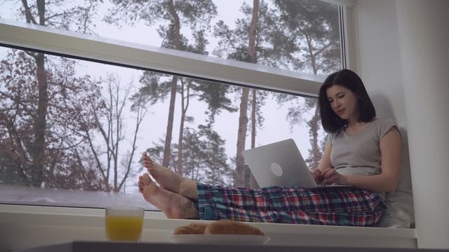 Girl working with computer sitting on windowsill. Casual woman wearing in t-shirt computing on pc in apartment with big panoramic windows. Beautiful winter landscape with snow and trees