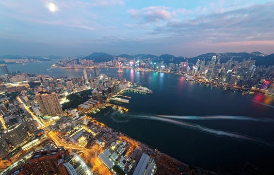 Aerial view of Hong Kong & Kowloon (Tsim Sha Tsui) at night with city skyline of crowded skyscrapers by Victoria Harbour & light trails of ships across seaport~ Cityscape of Hong Kong in blue twilight