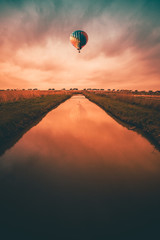Bunter Heißluftballon, der bei Sonnenuntergang über einen kleinen Fluss auf dem Land fährt.