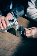 Close up of senior couple hands pouring coffee in a metal cup from thermos over a wooden table