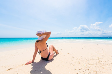 girl in hat and black swimsuit sitting on beautiful beach with blue ocean and sky on the background