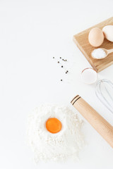 Cooking, culinary accessories and food ingredients lie on a white table. Eggs, flour, salt, pepper, whisk from the mixer, rolling pin and egg yolk. Space for text, daylight.