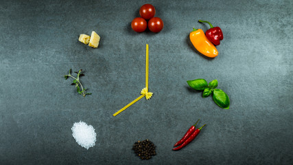 Clock out of pasta, tomatoes, paprika, basil, chili, pepper, sea salt, thyme and parmesan on dark stone background.